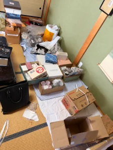 畳の部屋の壁際に箱入りの品、バッグ、小物、包装紙が置かれた様子